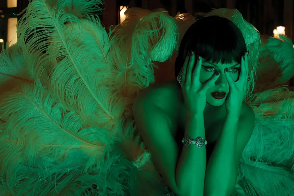 Person with dark hair and green lighting, surrounded by large feathers, holding their face with both hands