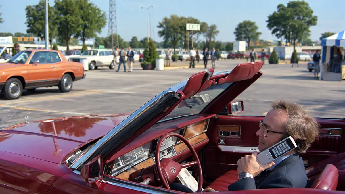 Man in a vintage red convertible using an early mobile phone in a parking lot with classic cars and people in the background